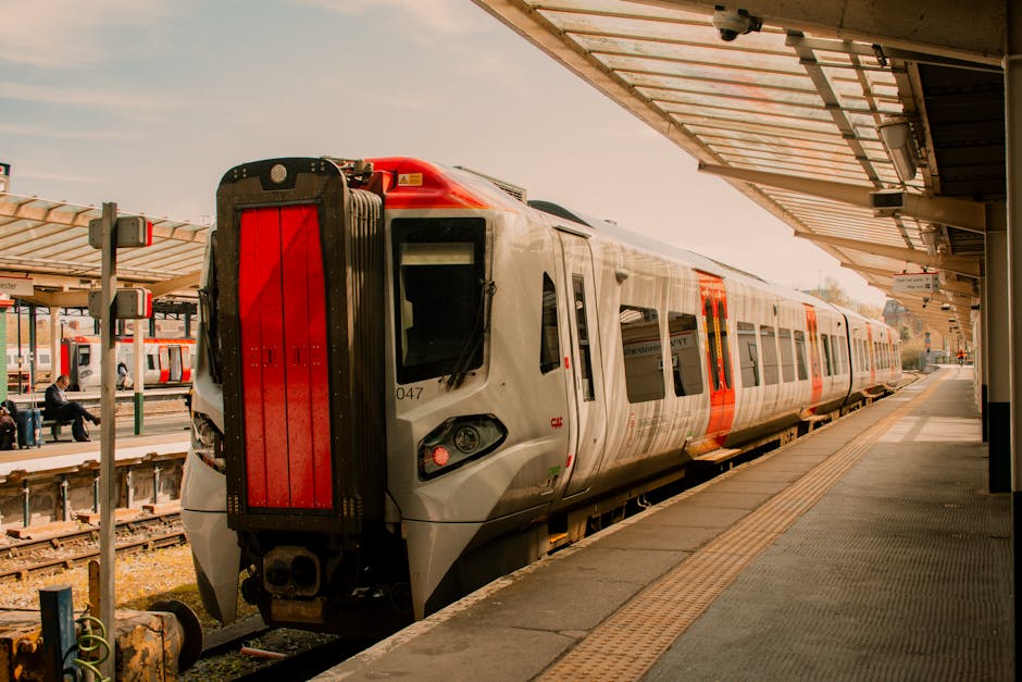 A modern electric train with a sleek, aerodynamic design is stationed at a railway platform, ready for boarding. The train's exterior features a silver body with prominent red accents along the sides,