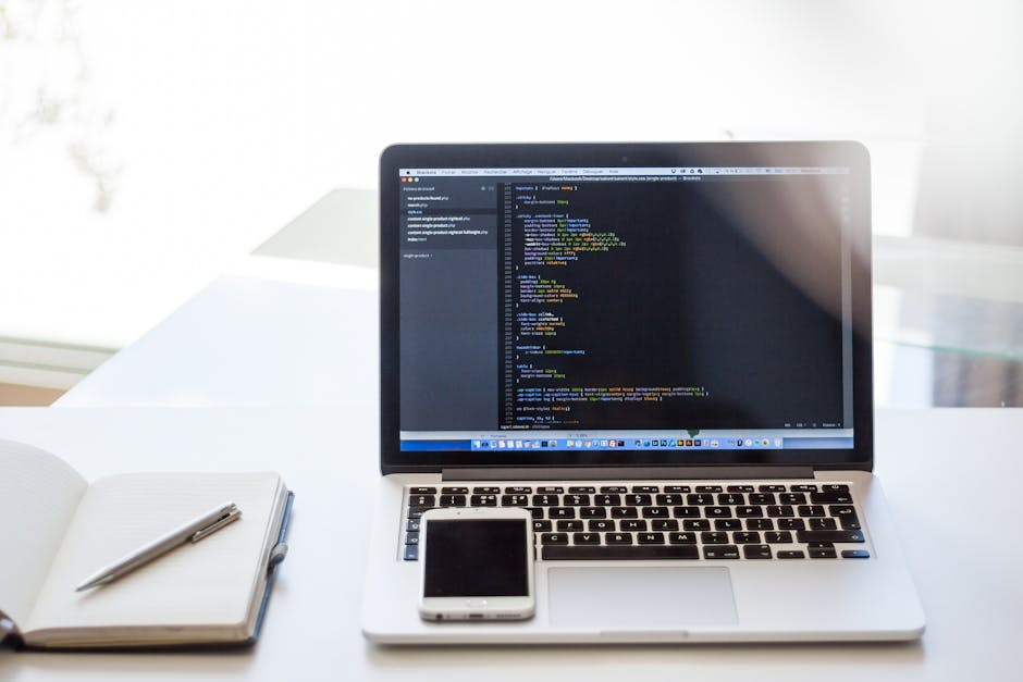 A white desk presented in a bright, well-lit room outdoors with a large window. On the desk, there is an open laptop displaying lines of colorful code on a dark background, indicating programming or w